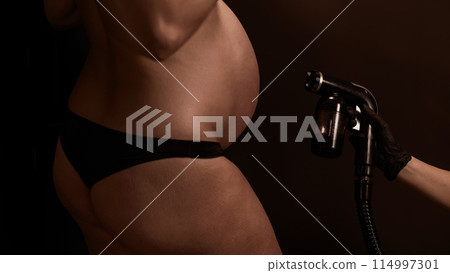 Close-up of the belly of a pregnant woman undergoing an instant tanning procedure. The master applies bronzer with a spray. Close-up of the belly of a pregnant woman undergoing an instant tanning procedure. The master applies bronzer with a spray. 114997301