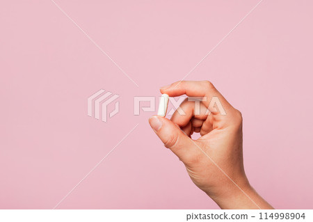 Woman hand holding a white pill against a plain pink background, copy space 114998904