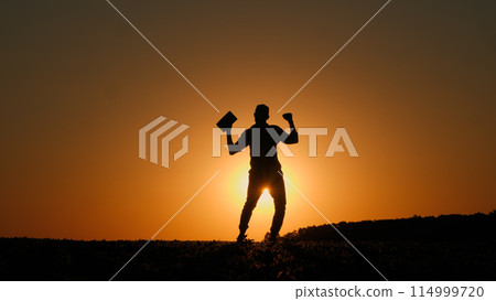 A man with a tablet in his hand dances a cheerful dance in a field at sunset 114999720