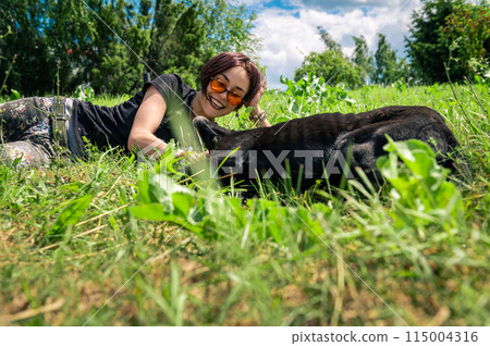 Beautiful happy girl lies and plays on the lawn with her dog. Young woman cuddling her dog on the grass. 115004316