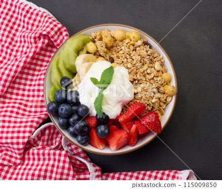 Healthy breakfast. Granola with strawberries, kiwi, banana and blueberries in a round plate on a black table. 115009850