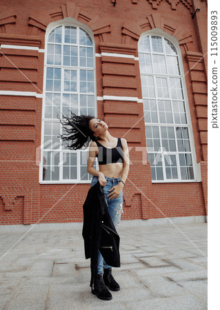 A woman in a black jacket and jeans poses in front of a brick building. Fashon street shot. A woman in a black jacket and jeans poses in front of a brick building. Fashon street shot. 115009893