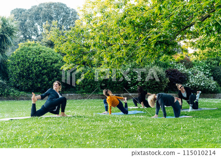 Female yoga class in park. Women stretching legs making low lunge or crescent pose exercising together with instructor on green grass lawn. Sport activity for fun, health, wellbeing, mental health. Female yoga class in park. Women stretching legs making low lunge or crescent pose exercising together with instructor on green grass lawn. Sport activity for fun, health, wellbeing, mental health. 115010214