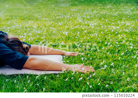 Cropped side view of woman doing yoga in Balasana or Child’s pose, breathing and meditation at summer park with green grass and blooming daisies. Sport activity for health, wellbeing, mental health. 115010224