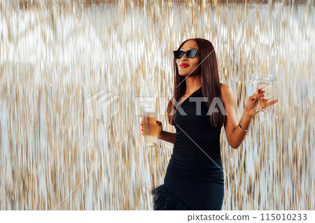 Young happy fashion African girl black woman in cocktail dress and sunglasses holding drink glass and wine bottle dancing at party with glitter background having fun at holiday, birthday celebration. 115010233
