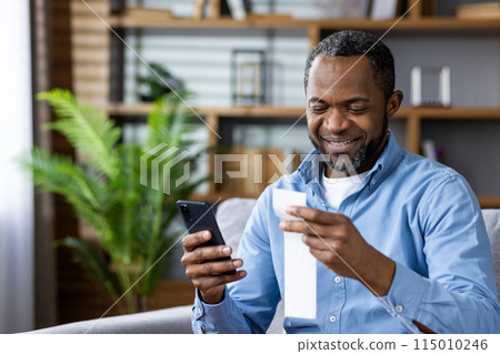 A man in his middle age is happily checking his smartphone, holding a receipt in a cozy home with a plant in the background. He seems content and engaged in modern technology 115010246