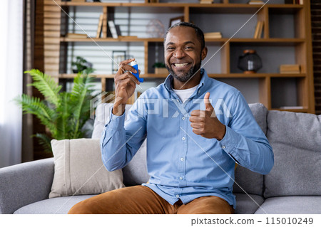 A cheerful man holding an inhaler and giving a thumbs up while sitting comfortably on a couch in a modern, well-decorated living room, emphasizing positive lifestyle and health. 115010249