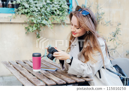 Happy smiling female traveler watching photos on digital camera while resting on cafe terrace with coffee in reusable cup after city walk. Travel blogger, photographer, influencer capture the moment. Happy smiling female traveler watching photos on digital camera while resting on cafe terrace with coffee in reusable cup after city walk. Travel blogger, photographer, influencer capture the moment. 115010250