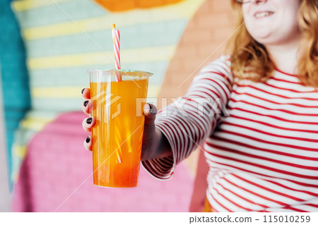 Glass of fruity sugar flavored tapioca pearl orange bubble tea with straw in hand of hipster fashion woman in bright clothes on the colorful painted brick wall background. Selective focus. Copy space. 115010259