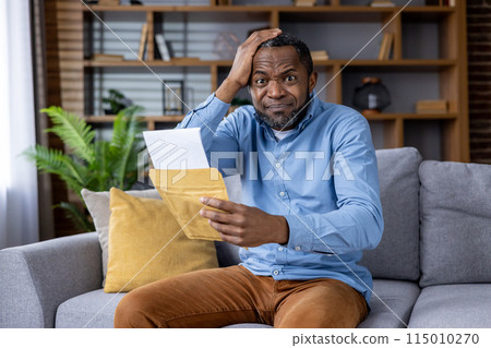 In a living room, a man sits on a couch, looking shocked as he reads a surprising letter. His body language reflects his emotional reaction to the unexpected news he just received at home In a living room, a man sits on a couch, looking shocked as he reads a surprising letter. His body language reflects his emotional reaction to the unexpected news he just received at home 115010270
