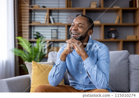 A happy person in a blue shirt laughing and relaxing on a cozy couch, surrounded by a stylish, modern living room with a bookshelf in the background and a green plant nearby. 115010274