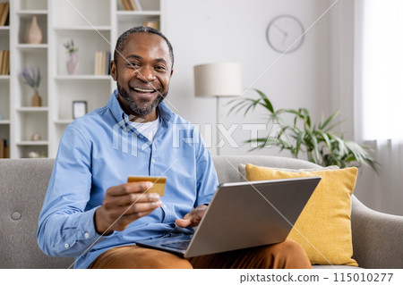 Smiling man holding a credit card, using laptop for online shopping at home. Comfortable living room setting with shelves and plant background. Lifestyle, technology, finance. 115010277