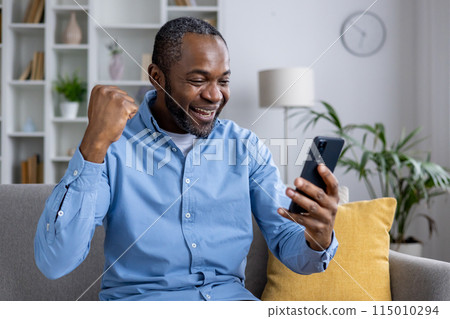 Joyful African American man in a blue shirt cheering with a clenched fist while looking at his smartphone in a cozy living room. Joyful African American man in a blue shirt cheering with a clenched fist while looking at his smartphone in a cozy living room. 115010294