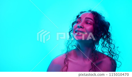 Happy, smiling, young woman with curly hair posing against cyan background in neon light. Spotless, well-kept skin 115010470