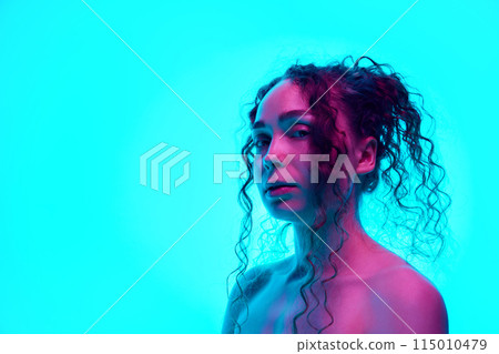 Portrait of beautiful young woman with curly hair in pony tail, well-kept skin, with bare shoulders posing against cyan background in neon light. 115010479