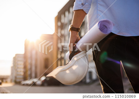 Unrecognisable engineer, architect or worker holds in hand protective white hard hat and blueprint on multi-apartment residential complex background, sunlighting. Concept of new real estate. 115010869