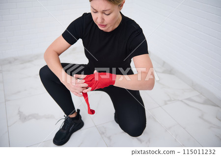 Determined confident European boxer fighter woman 40s, in black sports wear, tying tape around her hand before get boxing gloves preparing to boxing practice. Martial art and combat concept Determined confident European boxer fighter woman 40s, in black sports wear, tying tape around her hand before get boxing gloves preparing to boxing practice. Martial art and combat concept 115011232