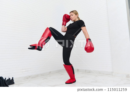 European blonde young kickboxing woman in black activewear and red kickboxing gloves, isolated white background performing martial arts kick. Sport exercise, fitness and workout. Full length portrait European blonde young kickboxing woman in black activewear and red kickboxing gloves, isolated white background performing martial arts kick. Sport exercise, fitness and workout. Full length portrait 115011259