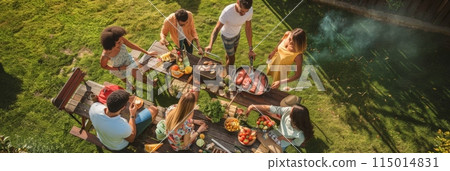 Aerial view of friends gathered around a summer barbecue table with grilled meats, salads, and drinks. Ideal for outdoor parties and social events. 115014831