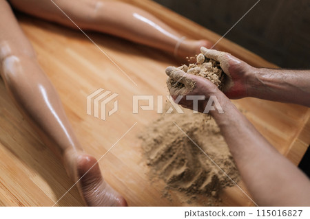 High-angle view of unrecognizable Ayurvedic massage practitioner male mixing heated sand on massage table for scrubbing treatment in luxury spa salon. Preparation to serenity of recreation 115016827