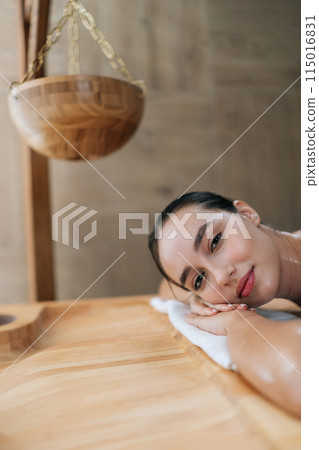 Vertical portrait of serene beautiful young woman lying relaxing on massage table looking at camera. Happy carefree woman enjoying leisure in spa parlor. Concept of healthy lifestyle and body care. Vertical portrait of serene beautiful young woman lying relaxing on massage table looking at camera. Happy carefree woman enjoying leisure in spa parlor. Concept of healthy lifestyle and body care. 115016831