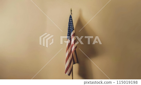 American flag on a stand with shadow on a beige wall. Simple and elegant patriotic display. American flag on a stand with shadow on a beige wall. Simple and elegant patriotic display. 115019198