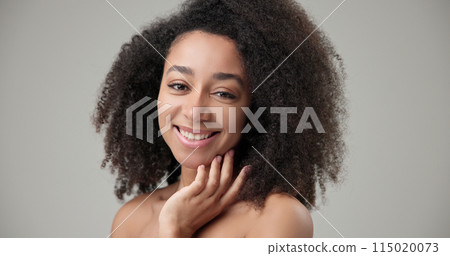 Beauty and healthcare concept - beautiful African American woman with curly afro hairstyle and clean, healthy skin touching face with hand, doing facial massage, posing and looking at camera Beauty and healthcare concept - beautiful African American woman with curly afro hairstyle and clean, healthy skin touching face with hand, doing facial massage, posing and looking at camera 115020073