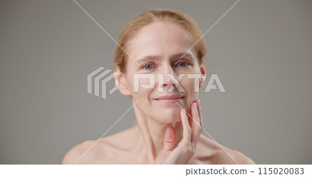 Anti age, Beauty, health and dry skin care concept - beautiful middle-aged mature Caucasian woman in her 50s touching her face skin and looking at the camera with a slight smile 115020083