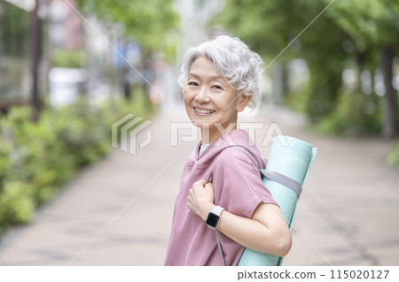 Senior woman holding yoga mat, looking at camera, smiling 115020127