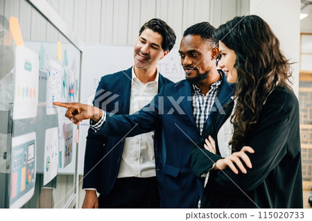 Business professionals in an office meeting use post-it notes on a glass board to brainstorm. Their diverse group emphasizes teamwork, leadership, and successful strategy discussions. 115020733