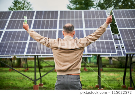 Back view of man happy about profit he got from investing in green energy. Man exulting with huge sum in hands and standing next to solar battery he bought. Back view of man happy about profit he got from investing in green energy. Man exulting with huge sum in hands and standing next to solar battery he bought. 115022572