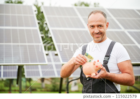 Knowledgeable man investing in green energy for future profit. Male adult happy about investment he did. Man in overalls putting euros to piggy bank on background of solar battery. 115022579