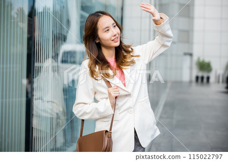 Portrait of confidence young businesswoman standing outside office building in city raise your hand to shade the sun. Happy woman wearing white suit jacket with brown bag at sunlight outdoors. 115022797