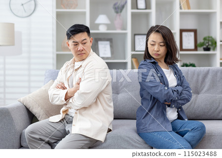 Couple in disagreement sitting apart on a sofa with arms crossed, depicting tension and unresolved conflict in a modern living room setting. 115023488