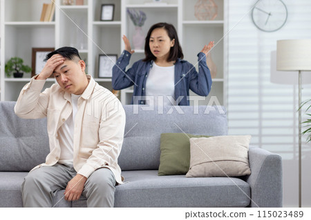 A stressed couple arguing at home in a modern living room. The man is sitting on the couch, while the woman stands behind with raised arms. A stressed couple arguing at home in a modern living room. The man is sitting on the couch, while the woman stands behind with raised arms. 115023489
