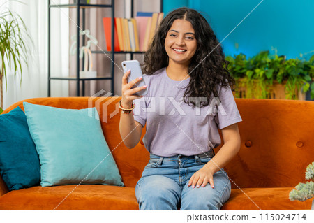 Indian Arabian young woman sitting on sofa use smartphone share messages on social media application 115024714