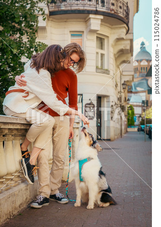 Young Caucasian couple sitting on curb and play with purebred dog Aussie. Vertical orientation. Pet training and city walking. Friendship with pet 115024786