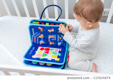 Little toddler girl playing with busy book while sitting in crib. Concept of quiet books and modern educational toys for kids Little toddler girl playing with busy book while sitting in crib. Concept of quiet books and modern educational toys for kids 115024874