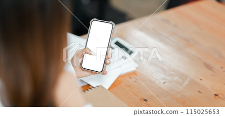 A woman is holding a cell phone with a white screen. She is sitting at a desk with a calculator and a notebook. The scene suggests that she is working or studying A woman is holding a cell phone with a white screen. She is sitting at a desk with a calculator and a notebook. The scene suggests that she is working or studying 115025653