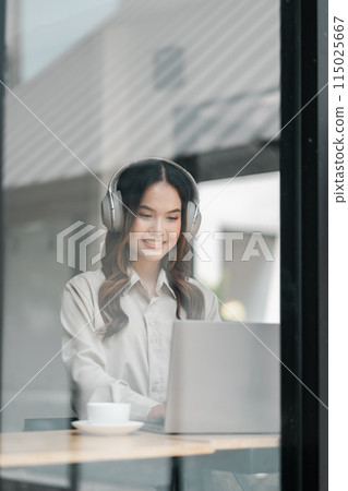 A woman wearing headphones is sitting at a table with a laptop and a cup of coffee. She is focused on her work, possibly listening to music or an audio recording A woman wearing headphones is sitting at a table with a laptop and a cup of coffee. She is focused on her work, possibly listening to music or an audio recording 115025667