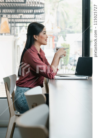 A woman is sitting at a table with a laptop and a cup of coffee. She is smiling and she is enjoying her coffee 115025727
