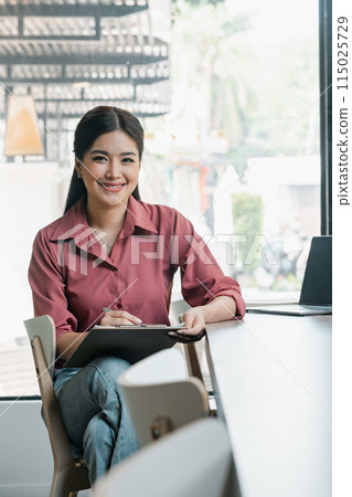 A woman is sitting at a table with a clipboard and pen. She is smiling and she is happy 115025729
