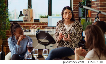 Asian woman entertaining friends at apartment party, sharing with them funny tale. Guest making multiethnic group of people laugh in living room while drinking alcoholic beverages Asian woman entertaining friends at apartment party, sharing with them funny tale. Guest making multiethnic group of people laugh in living room while drinking alcoholic beverages 115027101