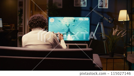Man in home theatre using cloud gaming service to play demanding science fiction videogame on TV. Gamer enjoying high quality graphics, streaming game over the internet, panning shot 115027146