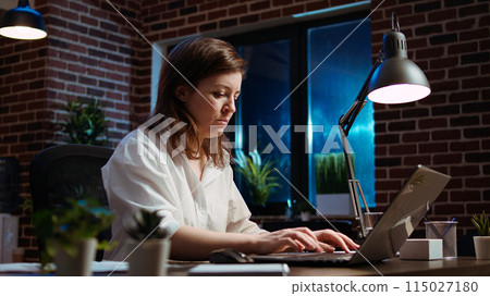 Businesswoman researching key data for company project, doing tasks in office overnight. Employee looking over accounting figures on laptop screen late at night, typing on keyboard, camera A 115027180