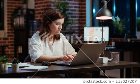 Office clerk looking through financial business documents on laptop late at night in workplace. Employee working on company project with deadline approaching soon during nightshift 115027207