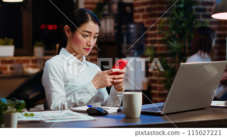 Smiling employee at work texting friends on cellphone, taking break from solving project tasks. Relaxed businesswoman using mobile phone to unwind in office late at night 115027221