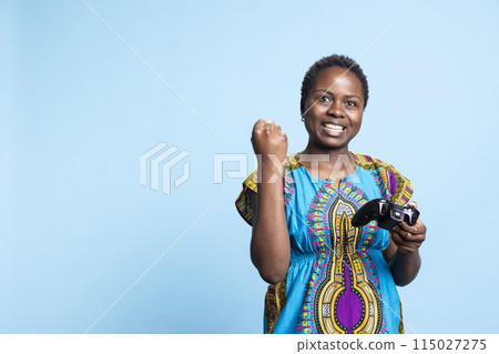 African american joyful girl winning videogames tournament in studio, having fun with gaming championship and playing with a controller. Female model being pleased with her gameplay victory. 115027275