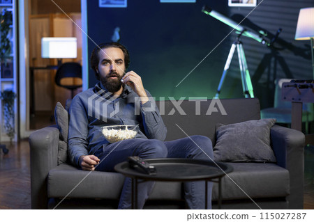 Man watching shows on TV and eating popcorn, engrossed by jaw dropping action, paying close attention to screen. Man sitting on couch in apartment eating popcorn, shocked by wild film plot 115027287