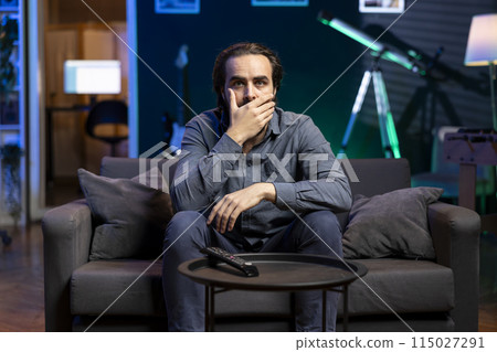 Viewer gasping while watching horror television series on streaming service, enjoying time off from work. Astonished person covering mouth in disbelief, shocked by film scene broadcasted on TV 115027291